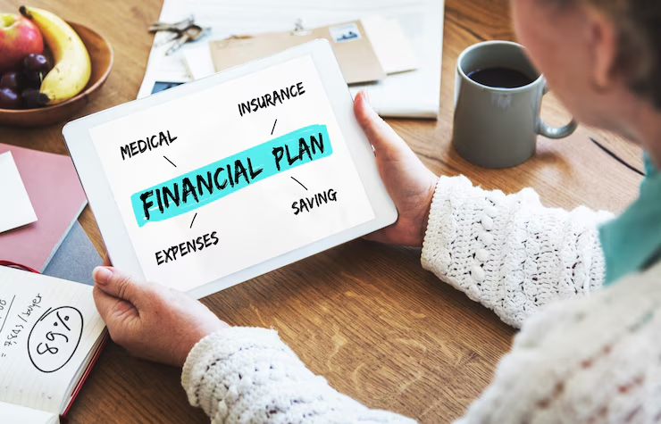 A person holding a tablet displaying a diagram titled "Financial Plan," with related terms branching out: Medical, Insurance, Saving, and Expenses. The tablet is placed on a wooden table next to a notebook, pen, and a gray coffee mug.