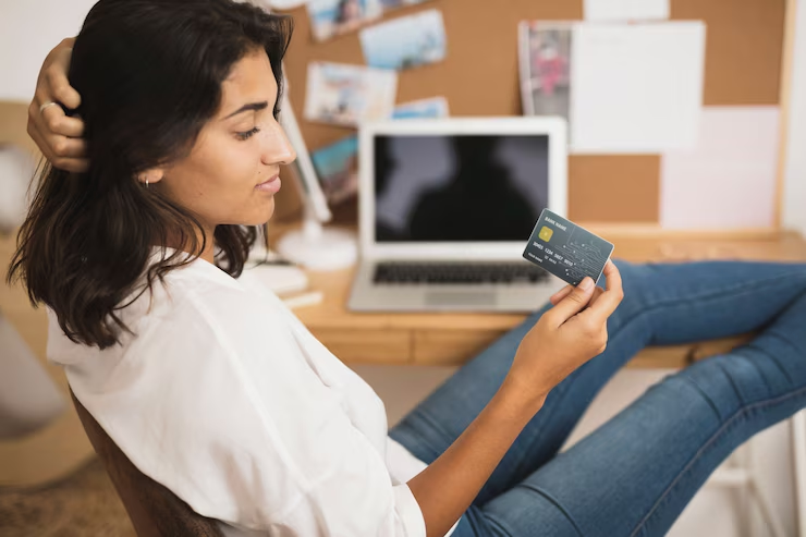 A relaxed young woman sitting at a desk with her feet up, holding a credit card and looking at it thoughtfully, with an open laptop and a corkboard in the background.