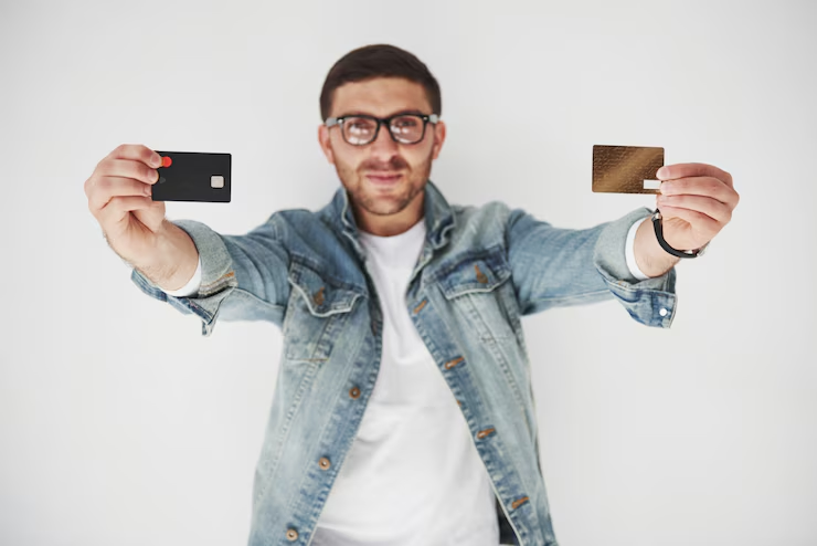 man wearing glasses and a denim jacket is holding out two credit cards—one black and one gold—toward the camera, smiling confidently against a plain white background.