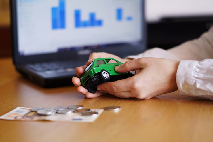 A person holding a green toy car near coins and banknotes on a desk, with a laptop displaying financial graphs in the background, symbolizing car financing, budgeting, or auto investment planning.