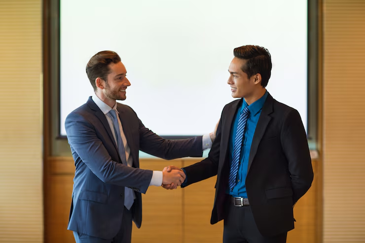 Two businessmen in formal attire are shaking hands and smiling in a professional setting, likely concluding a successful meeting or agreement, with a presentation screen in the background.