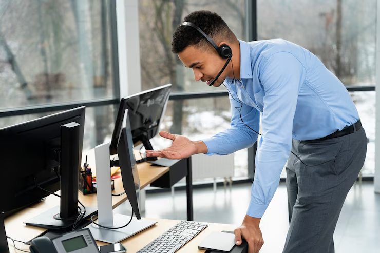 A customer service representative wearing a headset is standing and gesturing while speaking to a client through a video call or virtual meeting on a desktop computer in a modern office.








