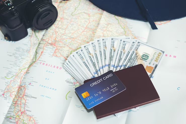 A travel-themed setup with a map of Thailand, a camera, a fan of hundred-dollar bills, a credit card, and a passport wallet, representing travel planning, finances, and budgeting.