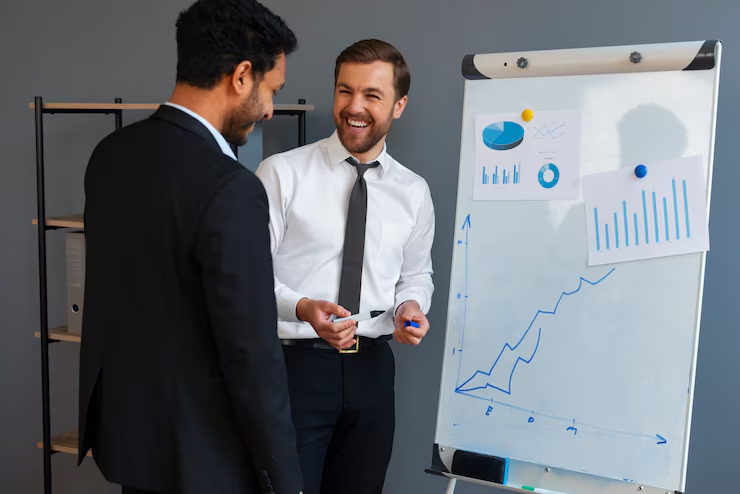 Two businessmen in formal attire are smiling and discussing data in front of a flip chart displaying graphs and charts, indicating a positive business presentation or strategy meeting.