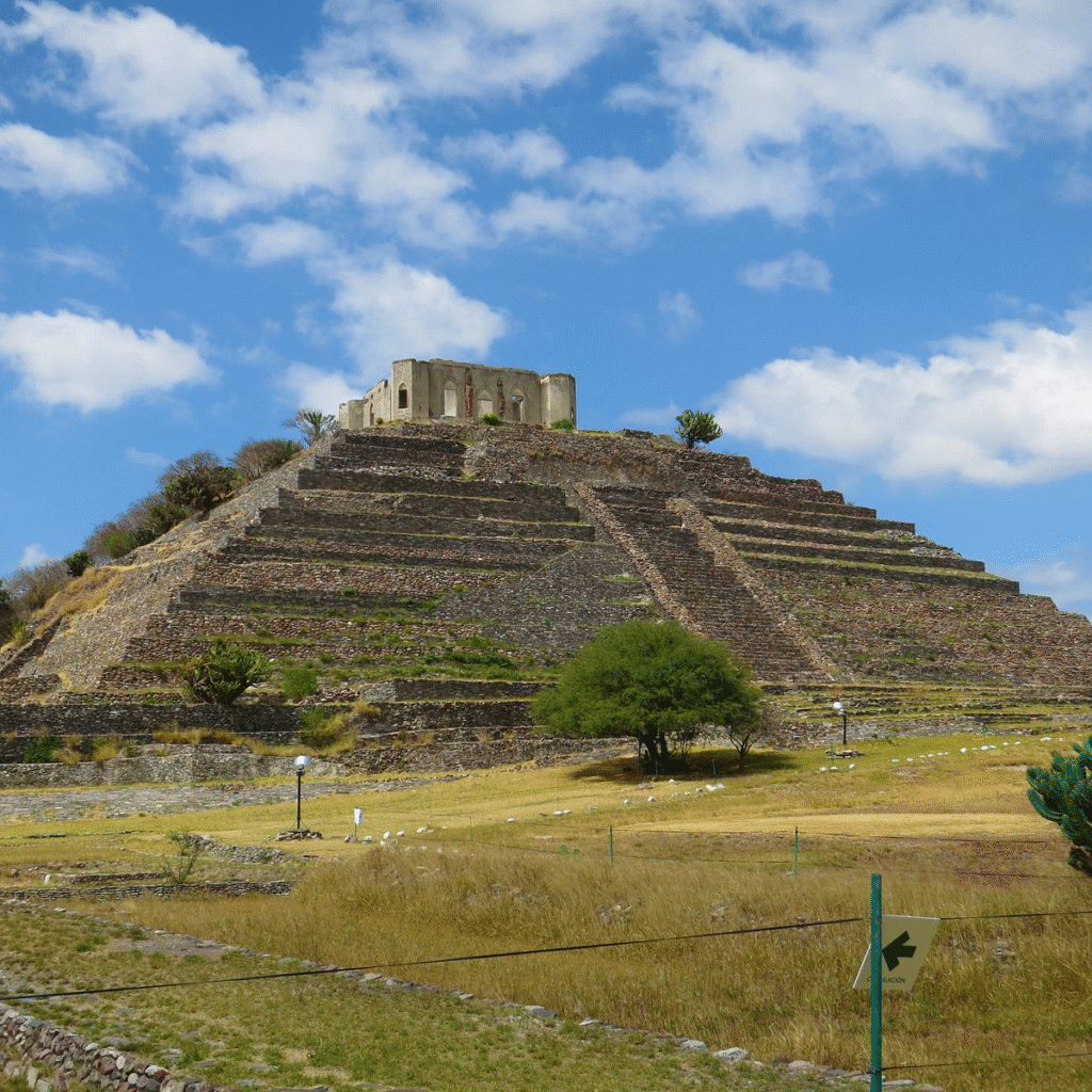 A large, ancient stone pyramid with a flat top and some ruined structures, under a blue sky with scattered white clouds, in a dry, grassy landscape.