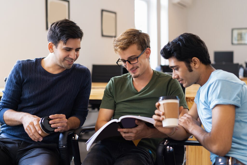 Three students reading textbook together and drinking from disposable cups. Young men sitting in classroom or library. Students concept. Front view.