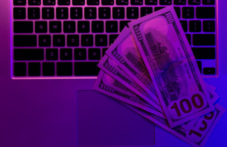A flat-lay photograph of a laptop keyboard with a stack of hundred-dollar bills on the touch-pad, illuminated by purple and pink light.