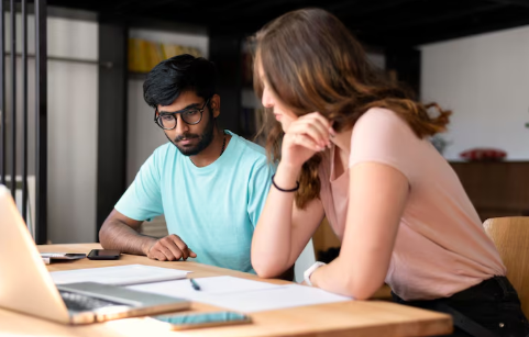 A male and female coworker are looking at paperwork and a laptop screen at a desk in an office.