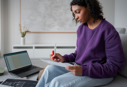 A woman sits on a couch, reviewing paperwork while a laptop with an open spreadsheet and floor plans is visible on a coffee table in front of her.
