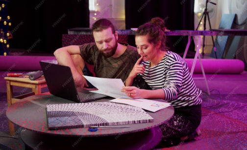 A man and woman kneel on a carpet, looking at a laptop and paper with a fan-shaped design, while working on an art or design project.