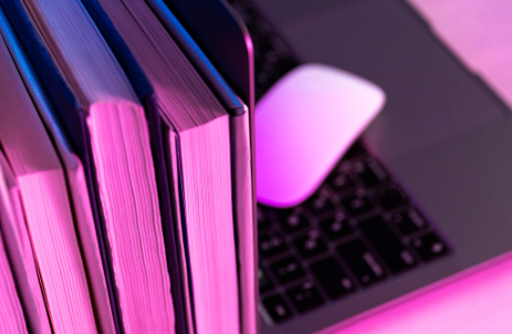 A row of hardcover books stands next to a white computer mouse on a laptop keyboard, all illuminated by a vibrant pink and purple light.