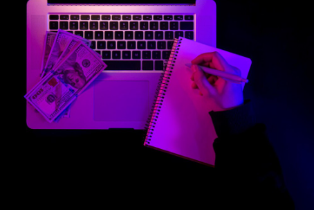 A hand writes on a notebook with a pen next to a laptop with a stack of hundred-dollar bills on the touch-pad, under a purple and blue light.