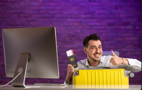A happy man with a yellow suitcase and a passport with plane tickets is sitting in front of a computer, giving a thumbs-up.