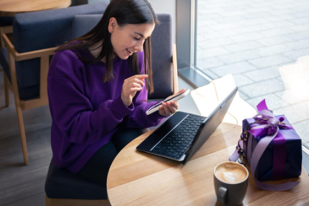 A woman in a purple sweater is sitting at a cafe table, using a smartphone and a laptop, with a coffee and a gift-wrapped box nearby.