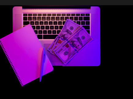 A person's hands are visible on a laptop keyboard. One hand is holding a stack of hundred-dollar bills while the other is in a fist, under purple and blue lighting.