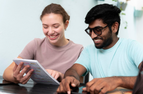 
A happy couple is laughing while looking at a sheet of paper with paint color swatches and holding a paint brush, while standing in a white-walled room.