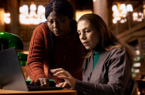 A person's hands are visible on a laptop keyboard. One hand is holding a stack of hundred-dollar bills while the other is in a fist, under purple and blue lighting.