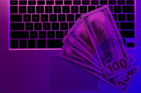 "Several $100 US dollar bills placed on a laptop keyboard, illuminated with purple lighting."