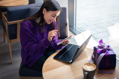 "Young woman in a purple sweater sitting at a café table, smiling at her phone while working on a laptop, with coffee and a gift box nearby."