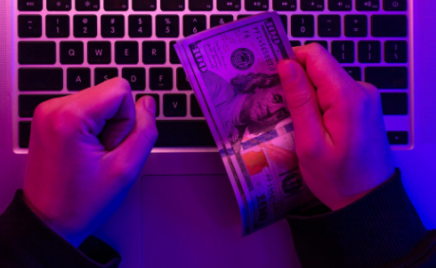 "Hands holding US $100 bills in front of a laptop keyboard under purple lighting."