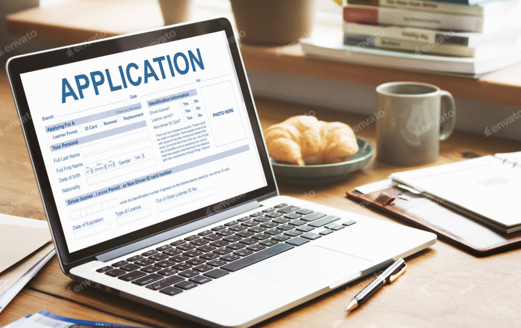 "Laptop on a desk displaying an online application form, with a notebook, pen, coffee mug, and croissant in the background."
