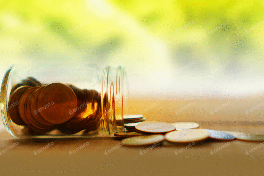 "Glass jar tipped over with coins spilling onto a wooden surface, with a blurred green background."