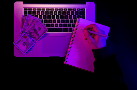 "Person writing in a notebook beside a laptop keyboard with several US $100 bills placed on it, under purple lighting."