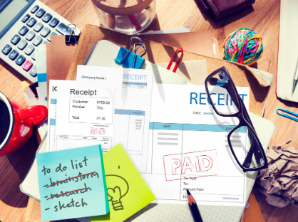 "Desk cluttered with receipts marked PAID, glasses, calculator, sticky notes, and office supplies."