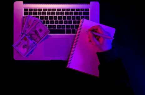 "Person writing in a notebook beside a laptop keyboard with several US $100 bills on it, under purple lighting."