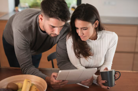 "Couple in a kitchen looking at a tablet together, with the woman holding a coffee mug and a fruit bowl on the counter."