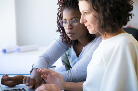 "Two women working together at a laptop, focusing on the screen and discussing something."