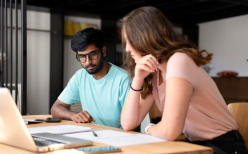 "Man and woman working together at a desk, looking at a laptop with papers spread out in front of them."