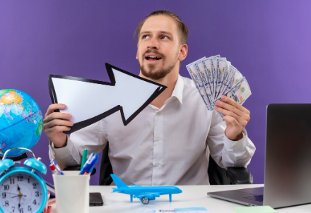 "Man sitting at a desk holding cash in one hand and a large arrow sign in the other, with travel items like a globe and toy airplane nearby."