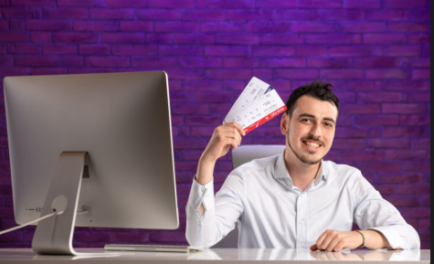 "Smiling man sitting at a desk holding airline tickets in hand next to a computer monitor, with a purple brick wall background."
