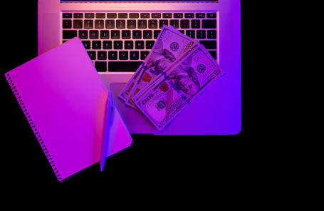 "Notebook and pen placed beside US $100 bills on a laptop keyboard, illuminated with purple lighting."