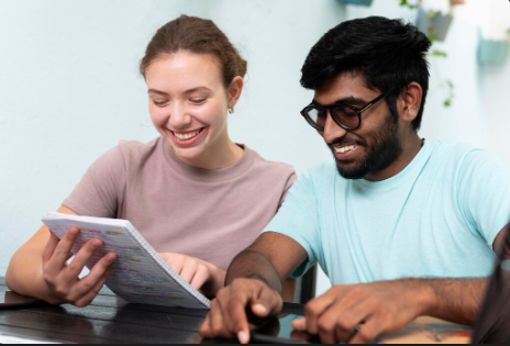 "Two students smiling while looking at notes in a notebook together at a table."