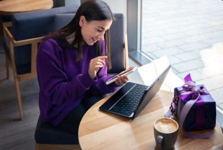 "Smiling woman in a café using her phone while working on a laptop, with a coffee and gift box on the table."
