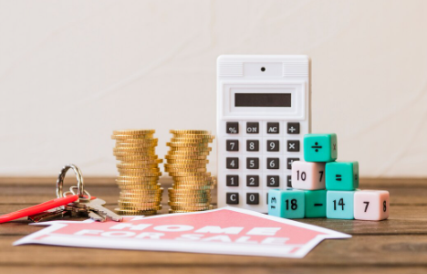"Stacks of coins, a calculator, house keys, and a 'Home For Sale' sign on a wooden table, symbolizing real estate investment."