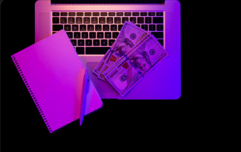 "Notebook with pen and US dollar bills placed on a laptop keyboard under purple lighting."