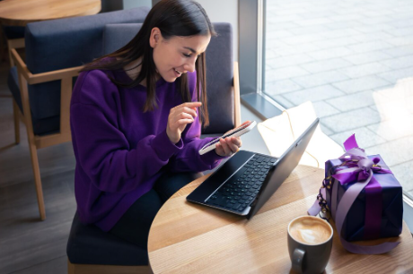 "Smiling woman in a purple sweater using her phone while working on a laptop at a café table with coffee and a gift box."