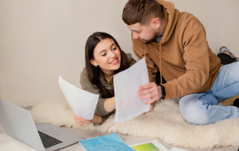"Smiling couple lying on a bed reviewing documents with a laptop and charts in front of them."
