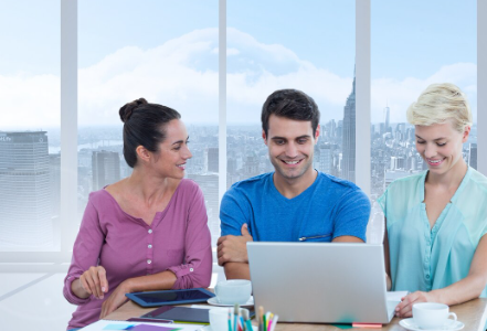 "Three colleagues smiling while working together on a laptop in a bright office with city view."